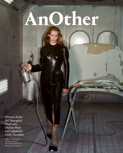 Woman in black latex outfit holding spray gun in rundown auto paint booth for high fashion magazine cover shoot with industrial vibe.