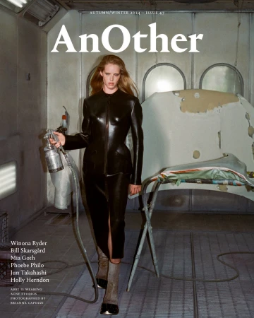 Woman in black latex outfit holding spray gun in rundown auto paint booth for high fashion magazine cover shoot with industrial vibe.