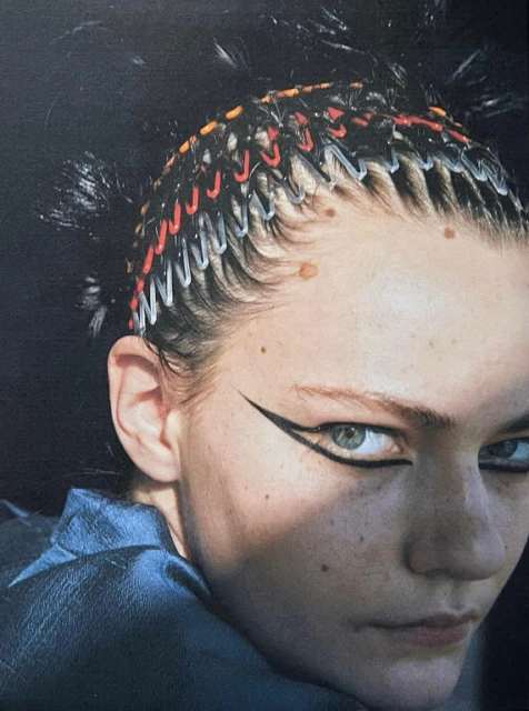 Close-up portrait of a young fair-skinned woman with graphic eyeliner and paper clip hair accessories against a dark background.