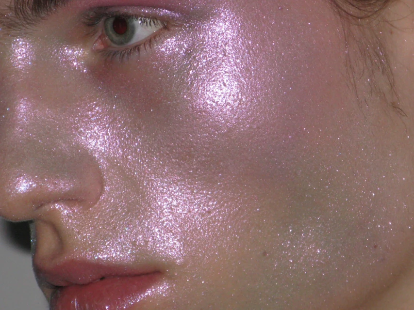 Close-up of a person's face with iridescent pink and purple makeup, emphasizing the dewy, shimmery skin and eye makeup details.