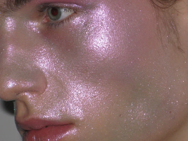 Close-up of a person's face with iridescent pink and purple makeup, emphasizing the dewy, shimmery skin and eye makeup details.