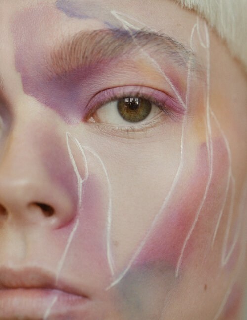 Close-up studio shot of a person's face with a neutral expression, featuring creative makeup, pink blush, and hand-drawn white outlines.