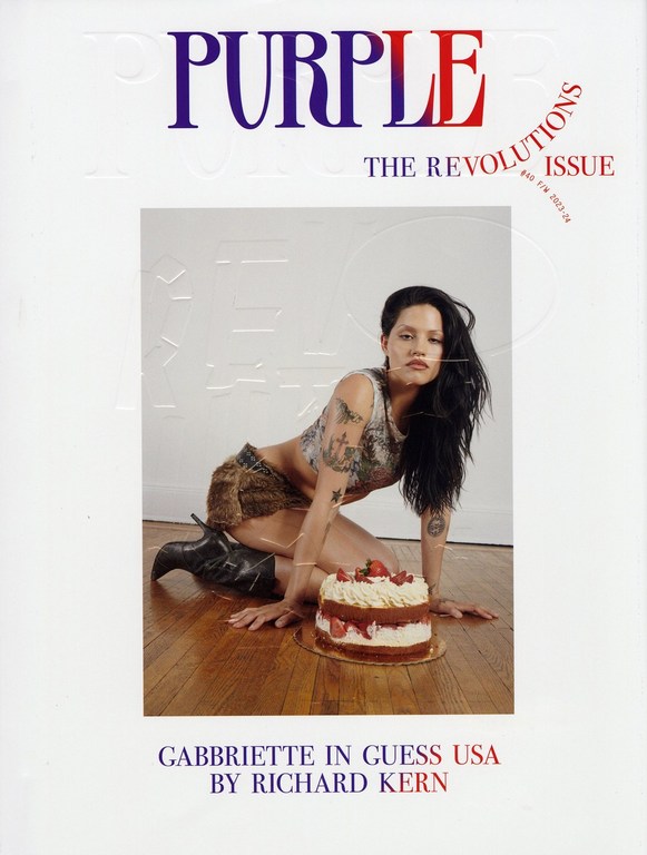 Gabbriette kneeling on wooden floor beside a strawberry cake wearing patterned crop top and dark boots, with purple magazine text above