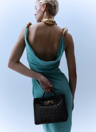 Model wearing a turquoise dress with a low back and gold collar, holding a dark woven handbag.
