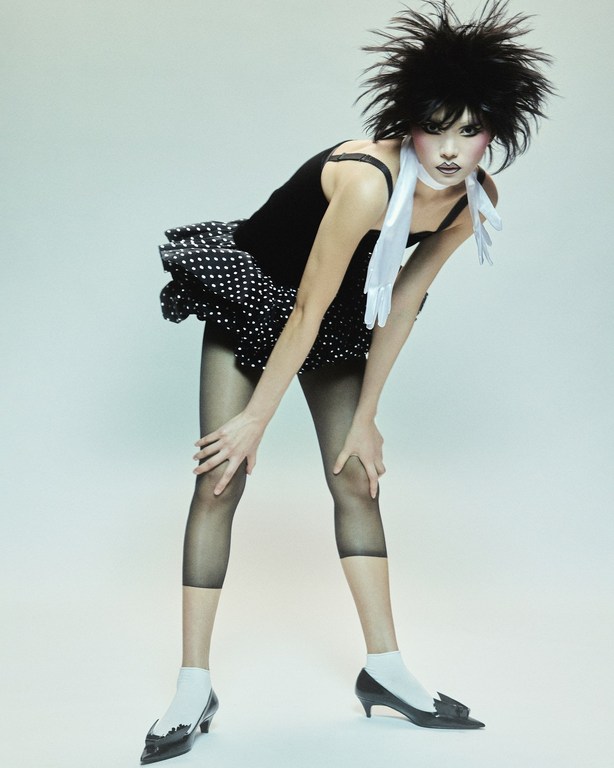A model poses in a black top and polka-dot skirt, captured in a studio setting with professional lighting, showcasing a fashion-forward look.