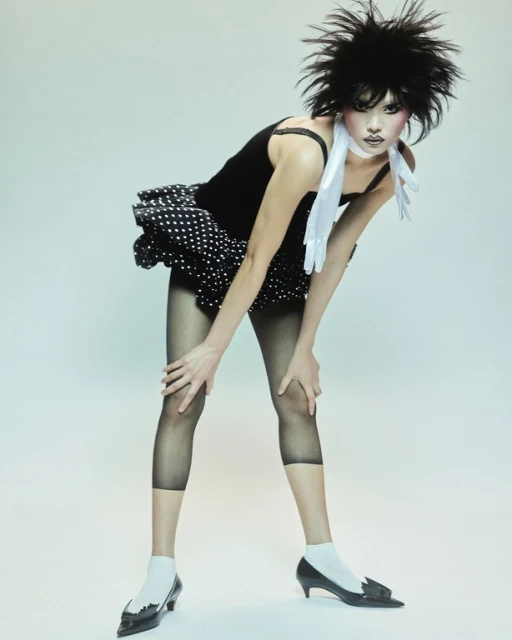 A model poses in a black top and polka-dot skirt, captured in a studio setting with professional lighting, showcasing a fashion-forward look.