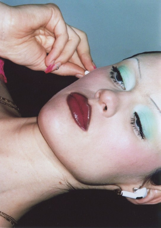 Close-up portrait of an adult female with an AirPod, pale skin, green eyeshadow, and dark red lips. Focus on beauty and personal technology.