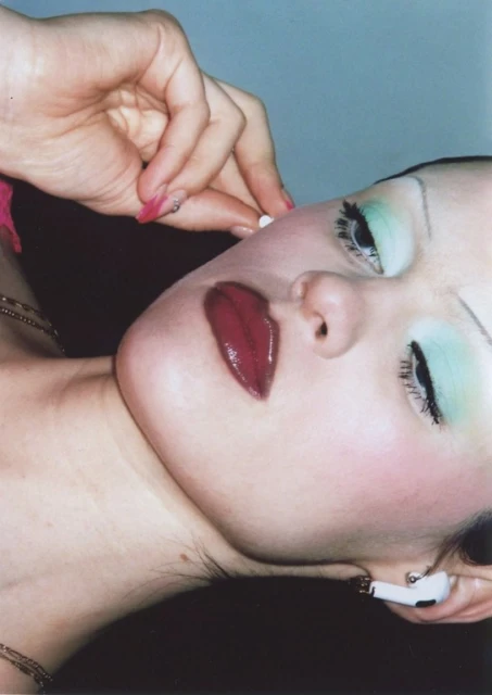 Close-up portrait of an adult female with an AirPod, pale skin, green eyeshadow, and dark red lips. Focus on beauty and personal technology.