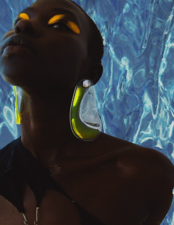 Portrait of a dark-skinned woman with yellow eye shadow and large, green earrings against a blue textured background.