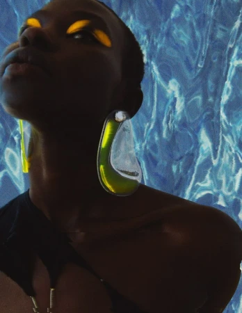Portrait of a dark-skinned woman with yellow eye shadow and large, green earrings against a blue textured background.