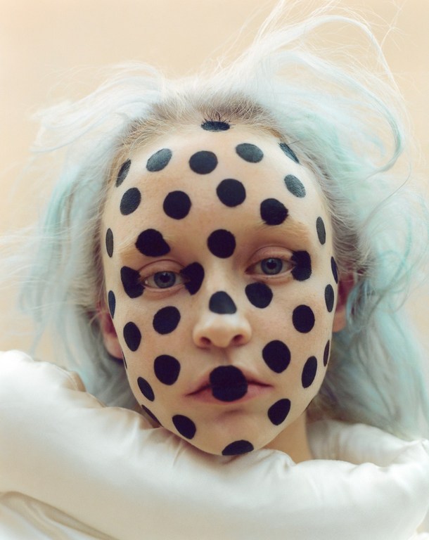 Close-up portrait of a young adult woman with a makeup of bold black dots covering her face and pale blue-tinged hair, against light background.