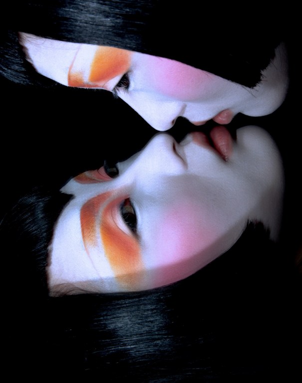 Close up portrait of two faces with pale complexions, dark hair, and striking orange and pink eye makeup reflected in a dark surface.