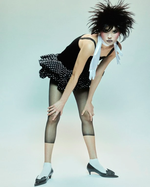 Model with spiky black hair and bold makeup wearing a polka-dot dress and sheer leggings on a gradient blue background