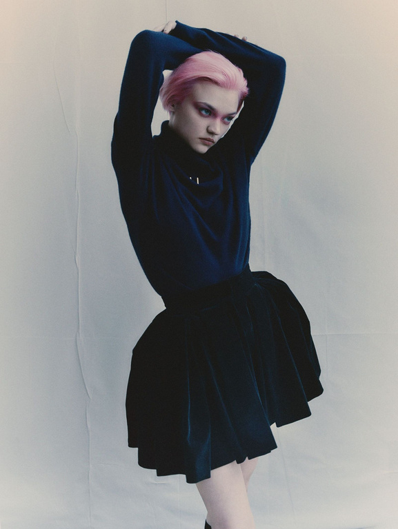 Young woman with pink hair and dark makeup wearing a black turtleneck and skirt against a white background in a fashion studio shoot.