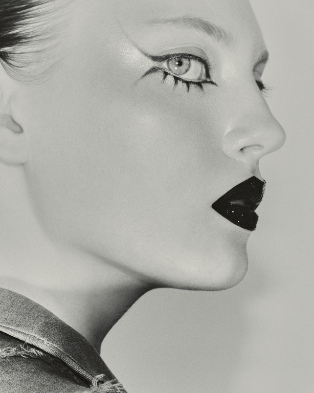 Close-up black and white portrait of a fair-skinned adult woman with dramatic black eye makeup, dark lipstick, and a denim collar.