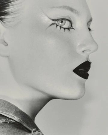 Close-up black and white portrait of a fair-skinned adult woman with dramatic black eye makeup, dark lipstick, and a denim collar.