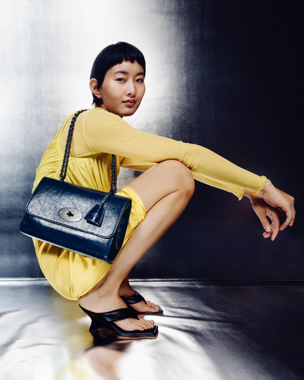 Woman in yellow dress squats with navy-blue leather handbag with chain strap in studio. She wears black heeled sandals.