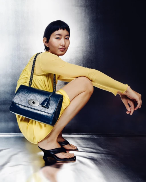 Woman in yellow dress squats with navy-blue leather handbag with chain strap in studio. She wears black heeled sandals.