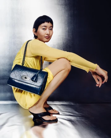 Woman in yellow dress squats with navy-blue leather handbag with chain strap in studio. She wears black heeled sandals.