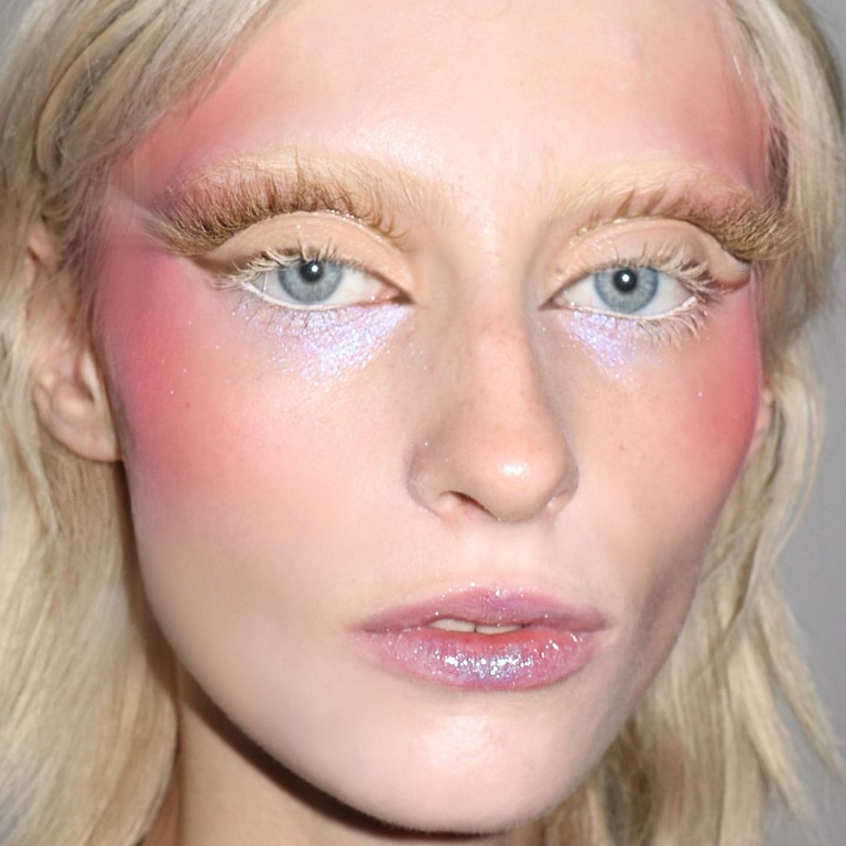 Close-up portrait of a woman with fair skin, light blonde hair, and striking eye makeup and blush in shades of pink and white.