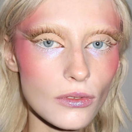 Close-up portrait of a woman with fair skin, light blonde hair, and striking eye makeup and blush in shades of pink and white.