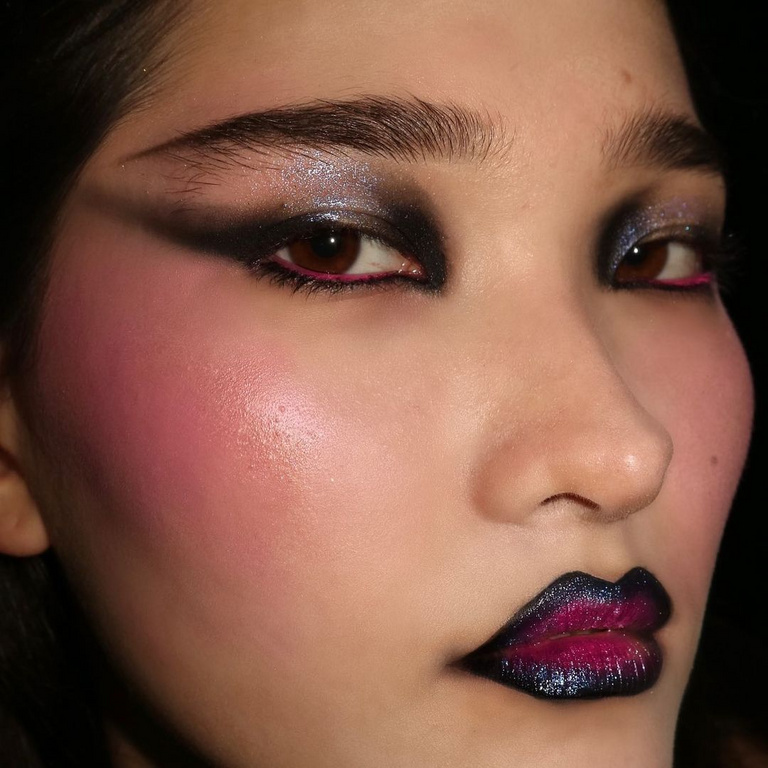 Close-up of a woman with dramatic makeup featuring black eyeshadow, bright pink blush, and dark iridescent lips on a neutral background.