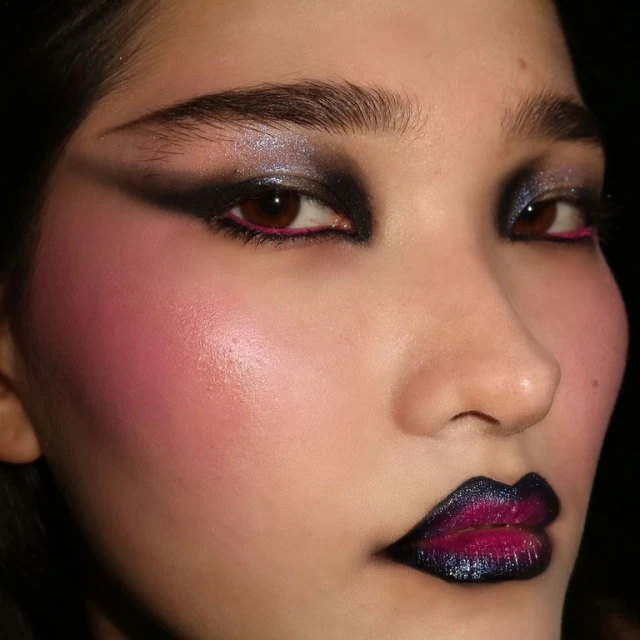 Close-up of a woman with dramatic makeup featuring black eyeshadow, bright pink blush, and dark iridescent lips on a neutral background.