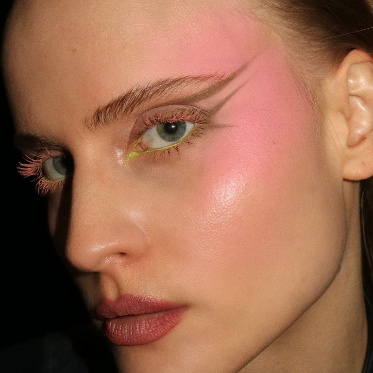 Close-up of a woman's face featuring pink blush and unique brown eyeliner art, combined with bright pink eyelashes. Beauty shot.
