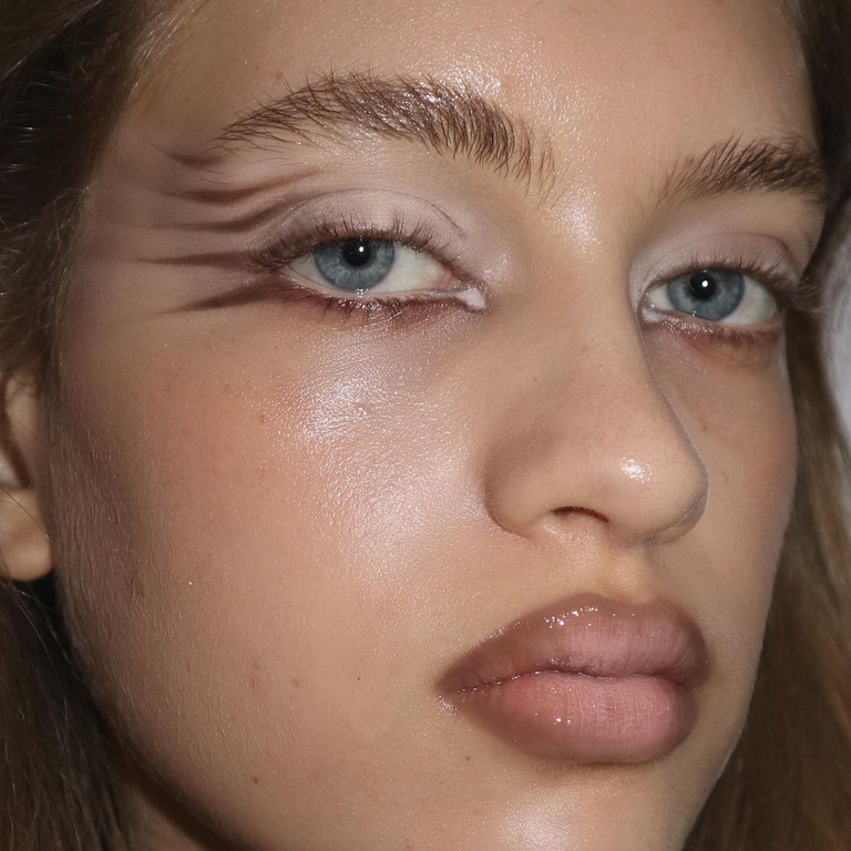 Close-up portrait of a light-skinned young woman with brown graphic eye makeup and glossy lips, highlighted features, and light blue eyes.