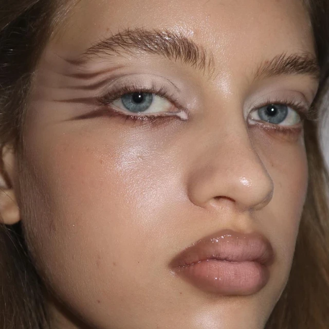Close-up portrait of a light-skinned young woman with brown graphic eye makeup and glossy lips, highlighted features, and light blue eyes.