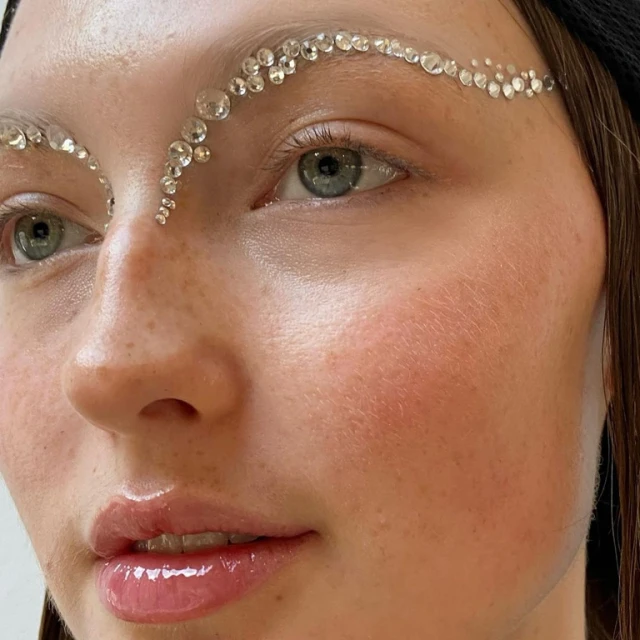 Close-up of a woman with clear gems above eyebrows and on her forehead, glossed lips, and wearing a black head covering. Beauty shot.