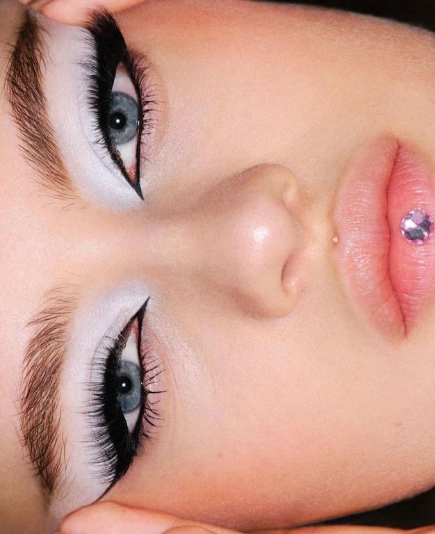 Close-up portrait of a white-skinned adult female with dramatic eye makeup, blue eyes, and a lip piercing with a gem.