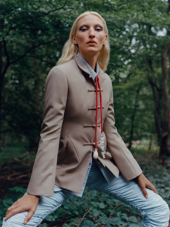 Fashion editorial shot of a blonde adult woman posing in a beige jacket and light blue patterned pants in a forest setting.