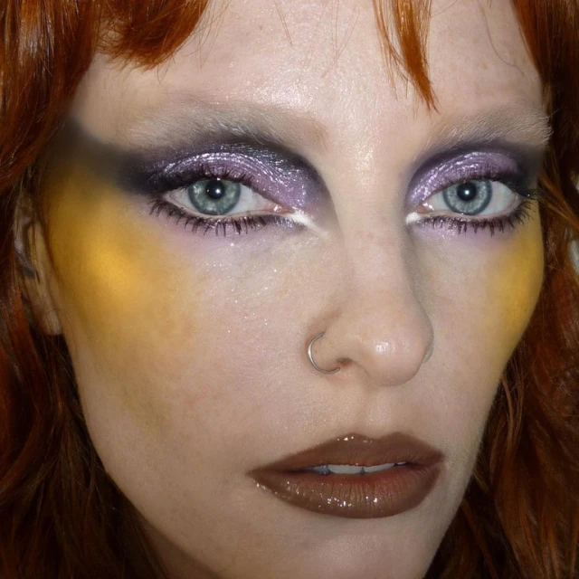 Close-up portrait of a woman with striking yellow and purple makeup, featuring glossy brown lips and a nose ring.