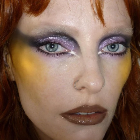 Close-up portrait of a woman with striking yellow and purple makeup, featuring glossy brown lips and a nose ring.