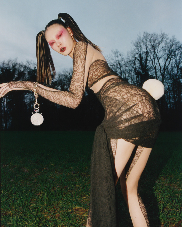 Woman in lace dress and bunny tail in outdoor fashion portrait, holding a pocket watch and posing near trees and green field.
