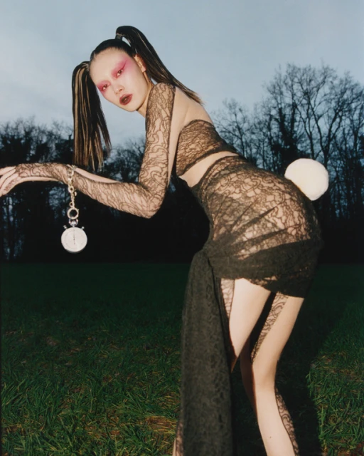 Woman in lace dress and bunny tail in outdoor fashion portrait, holding a pocket watch and posing near trees and green field.