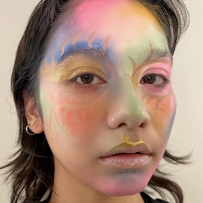 Close-up portrait of a person with vibrant, colorful artistic face paint in shades of pink, blue, green, and gold against a plain background.