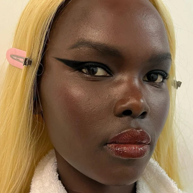 Close-up of a dark-skinned woman with blond hair, wearing black eyeliner and lip gloss, accessorized with hair clips, beauty shot.
