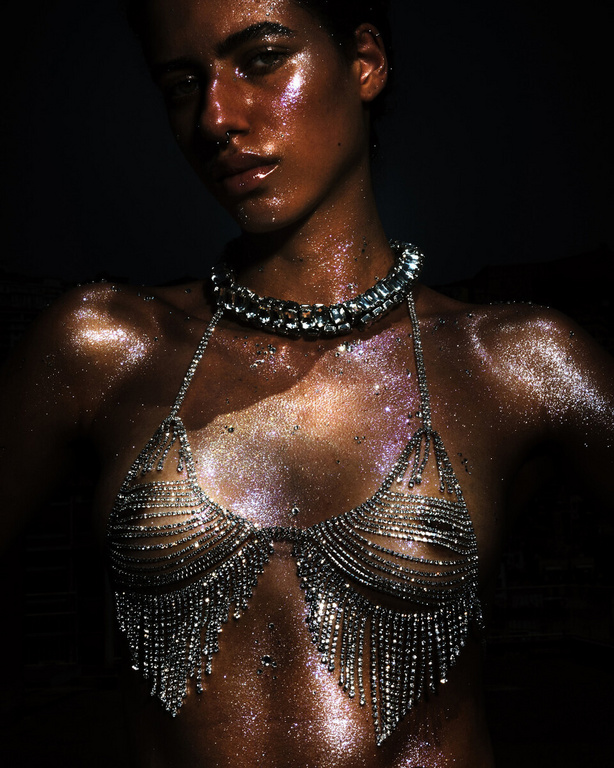 Glamorous adult female with glittery skin posing in jewelry-like top against dark background in indoor studio setting. Beauty, fashion.