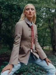 Portrait of a blonde woman wearing a tan jacket with knot closures and light blue pants outdoors in a forest.