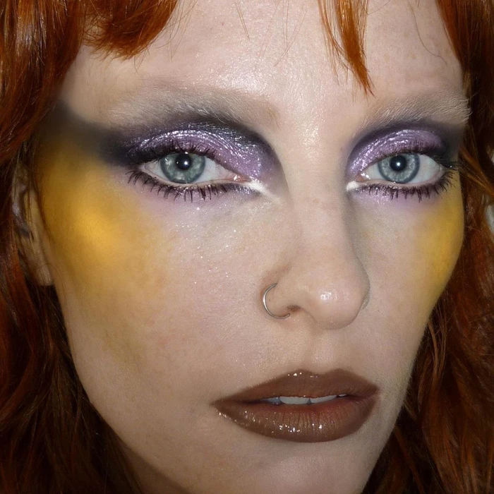 Close up beauty portrait of a woman with dramatic purple glitter eyeshadow, yellow blush, and glossy brown lips.