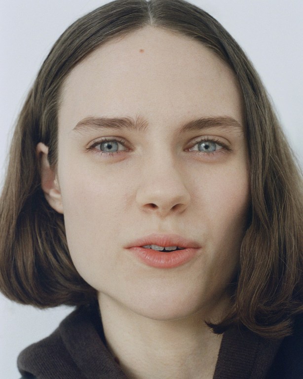 Close-up studio portrait of a blue-eyed, brown-haired adult woman with a neutral expression, wearing a brown top and set against a white backdrop.