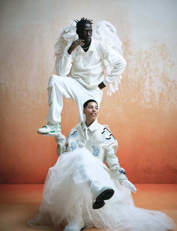 Two people in a studio with an orange background. One person is sitting wearing a white dress, another is sitting on top with wings.