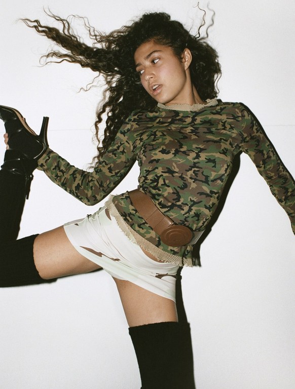 Young woman with curly hair posing in a camouflage top and black thigh-high boots against a white background in a studio shot.
