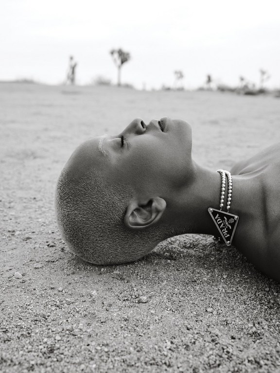 Close-up of a person lying on a sandy surface wearing a silver beaded necklace with a black Prada pendant in an arid landscape.