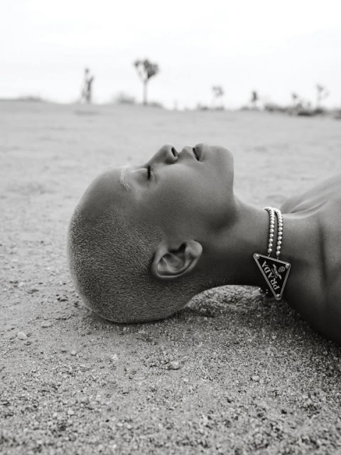 Close-up of a person lying on a sandy surface wearing a silver beaded necklace with a black Prada pendant in an arid landscape.