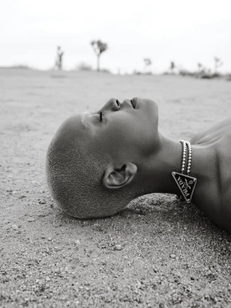 Close-up of a person lying on a sandy surface wearing a silver beaded necklace with a black Prada pendant in an arid landscape.
