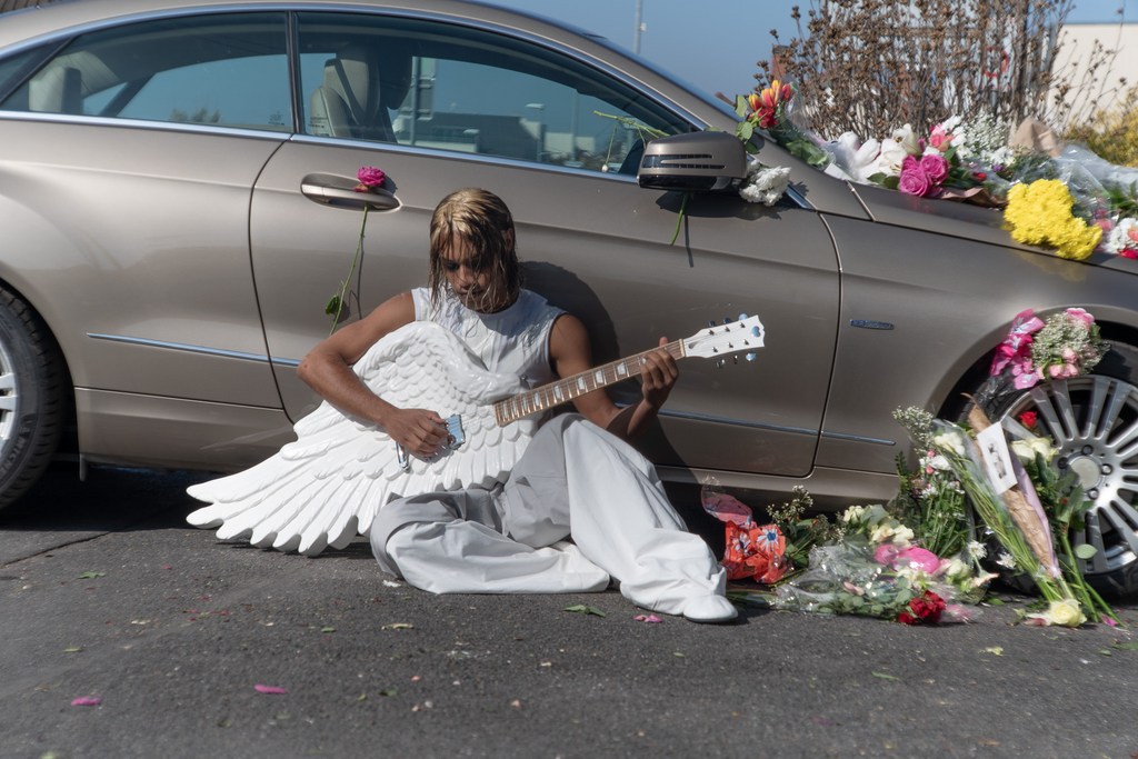 Man with angel wings playing guitar next to gold car decorated with flowers in outdoor setting on asphalt road.