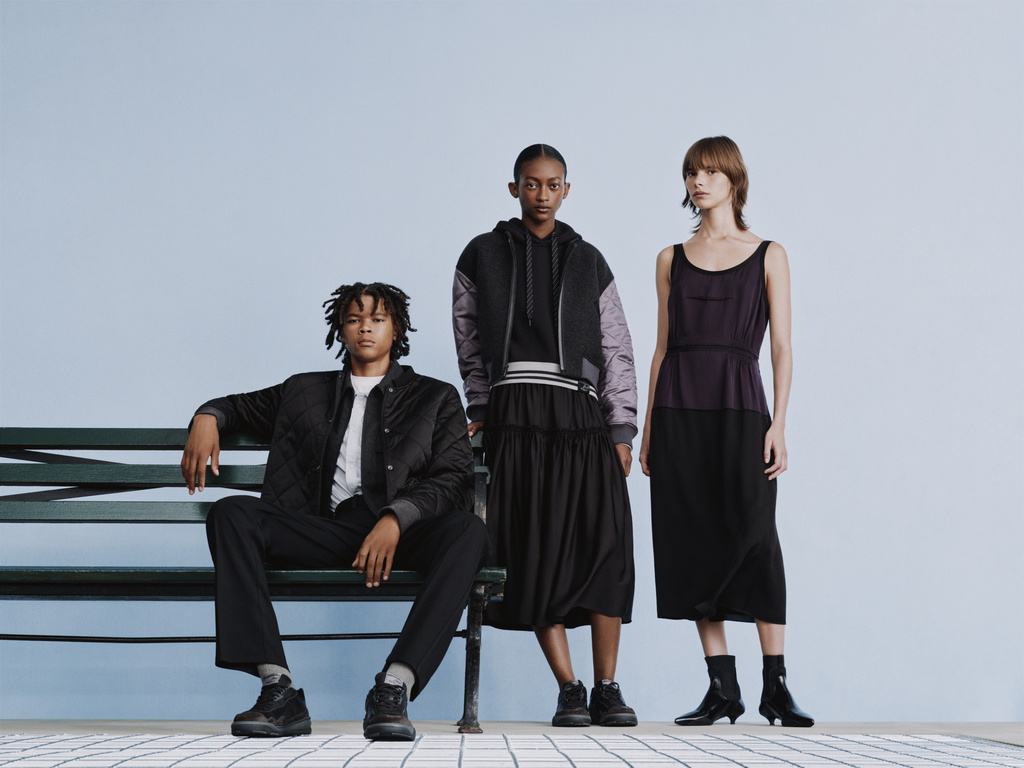 Three fashion models pose in black and grey clothing on a bench against a light blue backdrop in a studio setting.